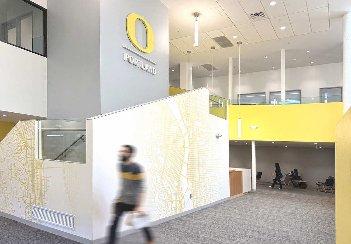 The interior lobby of the UO Portland Campus Center features yellow wall graphics represesnting an aerial map of Portland. A large "O" for the University of Oregon mounted above the stairwell.