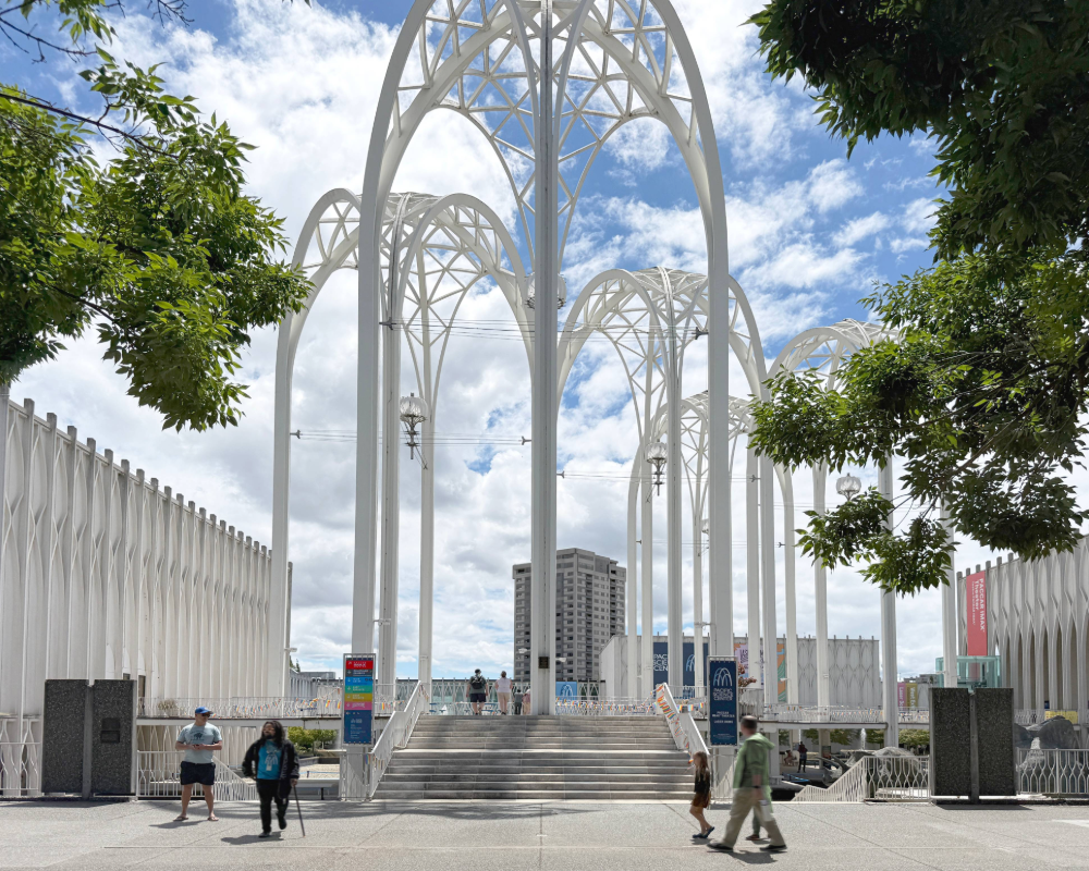 The newly opened courtyard at Pacific Science Center, framed by the historic 1962 World's Fair arches. Wayfinding banners with maps and directional signage are mounted at the entrance to the courtyard stairs.
