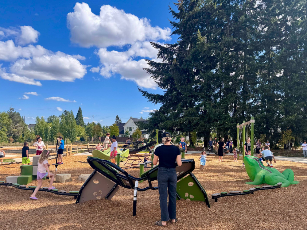 Beneath a swatch of mature evergreen trees, children play on the new, bright green playground equipment at Frog Pond Park. A concrete frog sits proudly amongst the play equipment, both as the park's mascot and a place to play and climb.