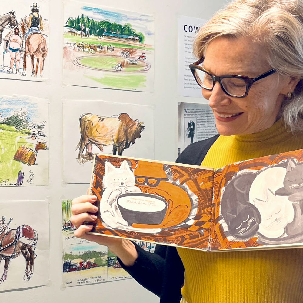 Debbie Shaw holds up a book illustrated by Chuck Hayden, standing in front of his sketches hung on the M/R Gallery wall.