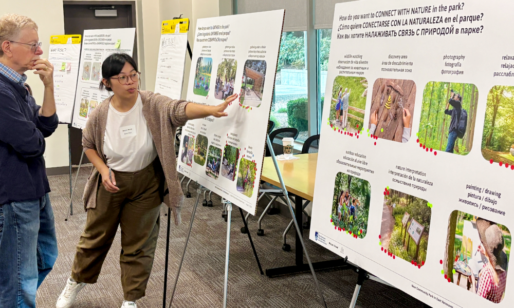 A Mayer/Reed landscape architect stands with an east Vancouver community member, pointing at the large multilingual display boards at a community engagement event. The boards feature images different types of programming and park uses. Participants marked images they are drawn to with red dots.